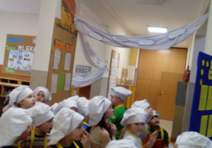 Dzieci w strojach kucharzy podczas zwiedzania wystawy w przedszkolnym holu.
